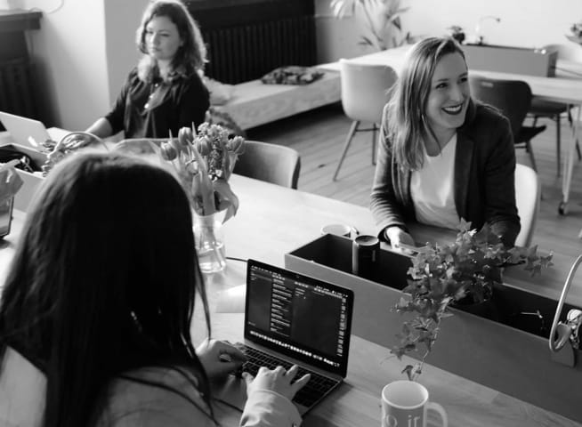 A picture displaying three persons sitting in an office environment. One of them is smiling and talking to someone outside of photo's frame, other two are working with code behind their laptops.